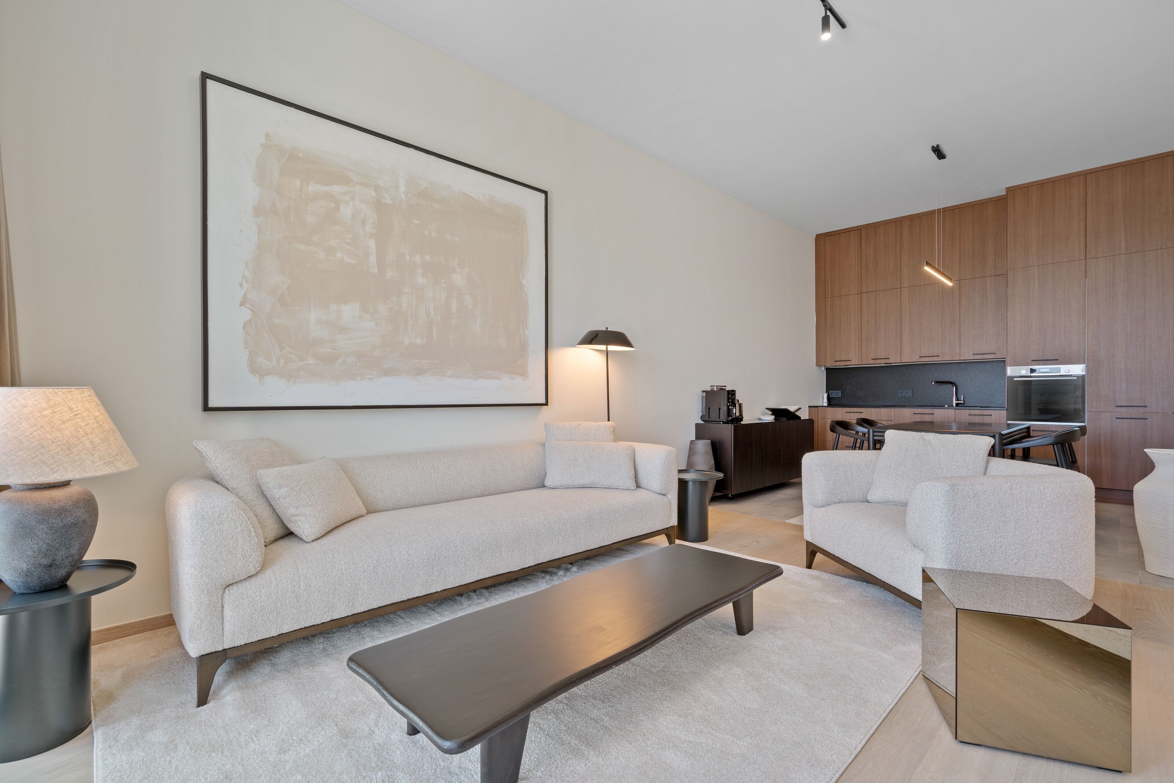 A modern living room with a sofa, a coffee table, and a lamp. There is a kitchen area with cabinets and appliances in the background.