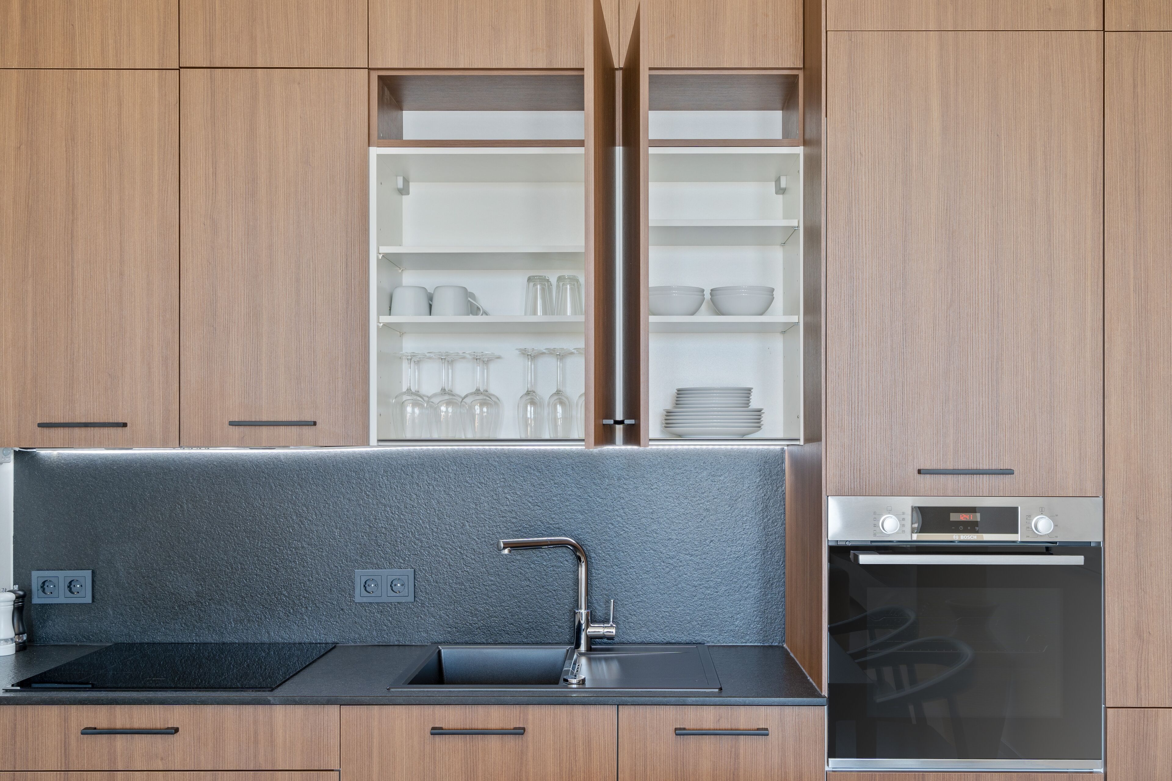 A modern kitchen with wooden cabinets, a black countertop, a built-in oven, and a glass-fronted shelving unit with dishes and glasses.
