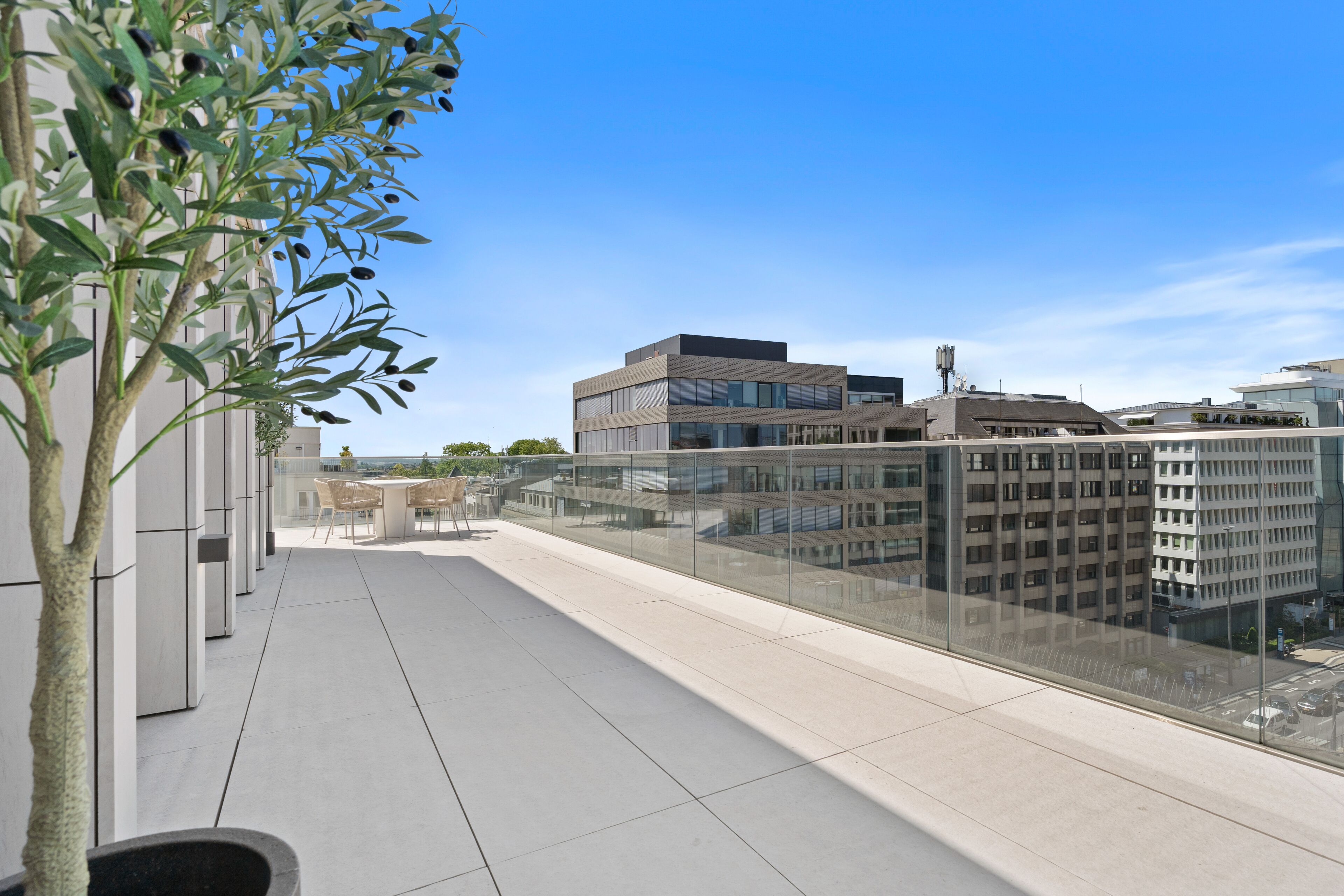 A modern building with a glass facade, a flat roof terrace with chairs, and a view of other buildings.