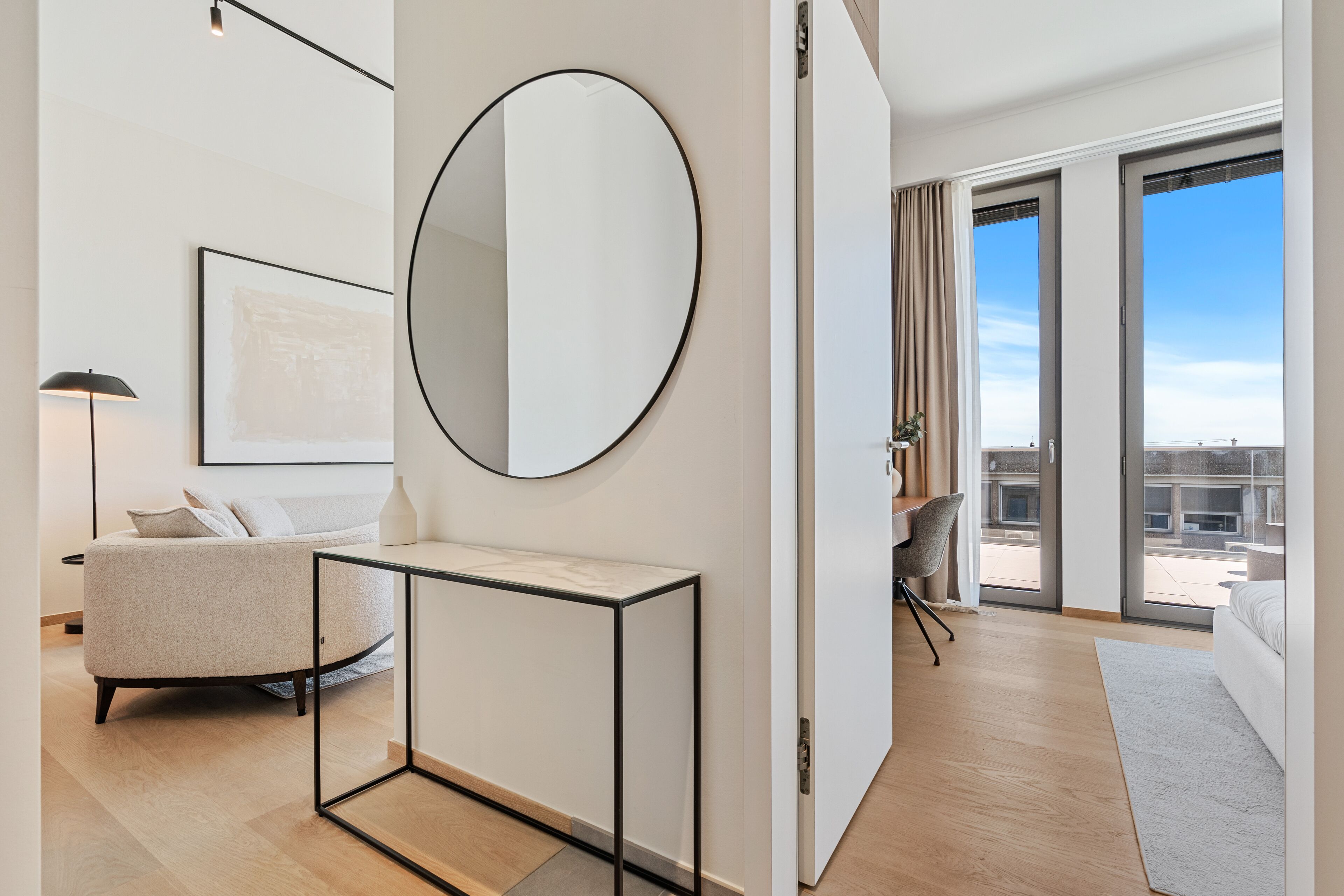 Ein modernes Hotelzimmer mit großem Fenster, einem runden Spiegel, einer Sitzecke, einem Schreibtisch und einem Sessel.