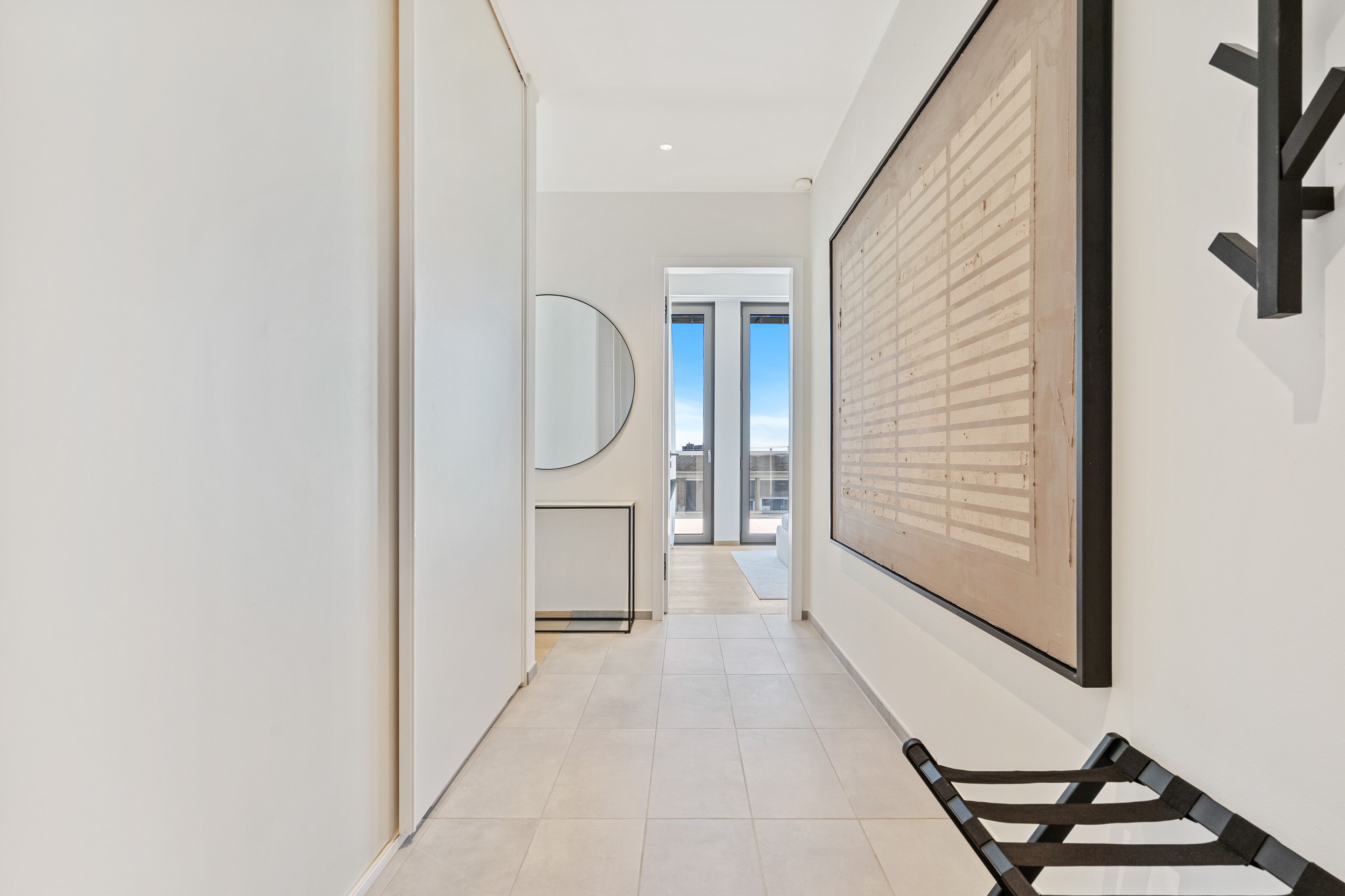 A hallway with a large framed artwork on the wall, a mirror, and a view of the outdoors through a door.