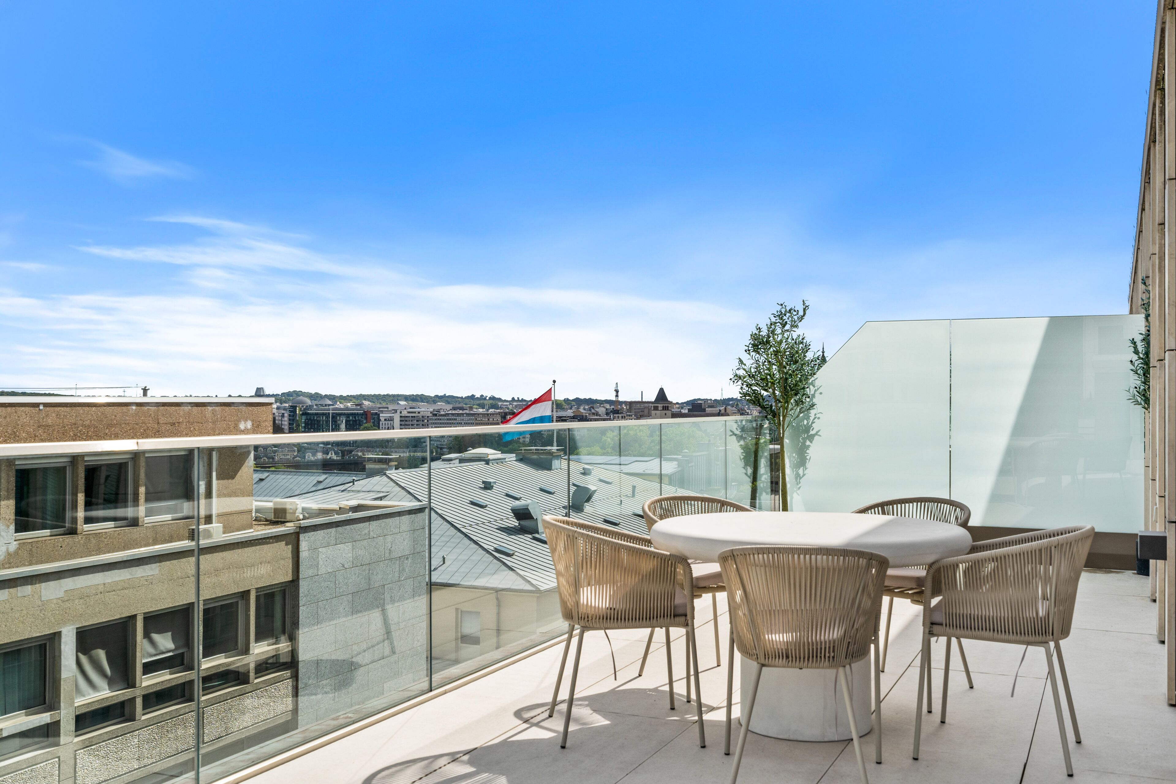 A rooftop terrace with a glass balustrade, a round table with chairs, and a view of the cityscape.