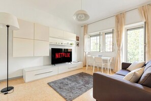 A modern living room with a flat-screen TV, white cabinetry, a grey rug, a brown sofa, and a dining area with white chairs.