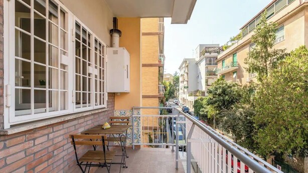 Un balcón con un muro de ladrillo, una ventana con marcos blancos, una mesa con sillas y una barandilla de metal.
