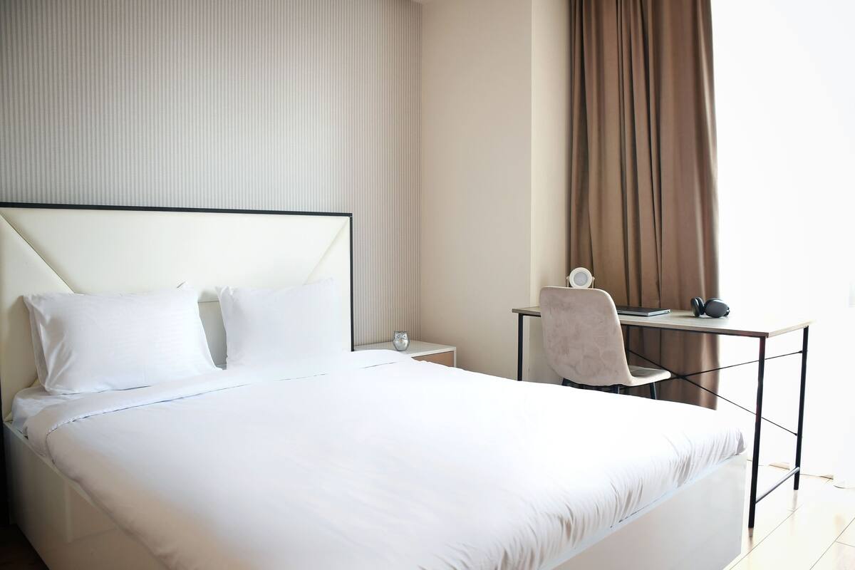 A hotel room with a large bed, a desk with a chair, a small table, and a window with curtains.