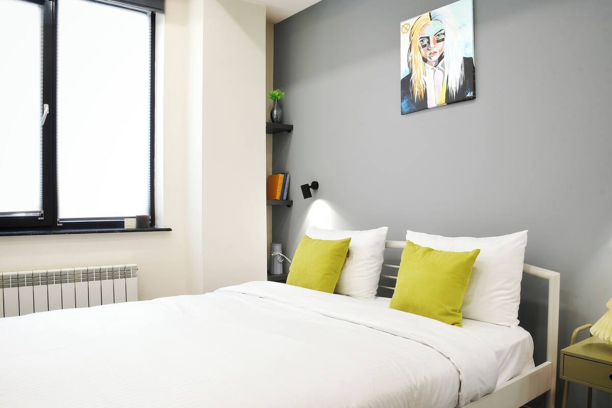 A modern bedroom with a large bed, a window, a radiator, a bedside table, and a wall-mounted shelf with books and a lamp.