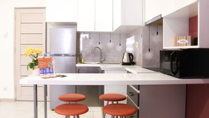 A modern kitchen with white cabinets, a stainless steel sink, a kettle, and a microwave. There are two orange stools at the counter.