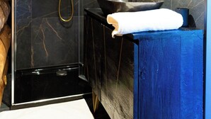 Een moderne badkamer met een zwarte marmeren wand, een houten plafond, een glazen douchecabine, een wastafel met een blauwe onderkast en een ronde spiegel.