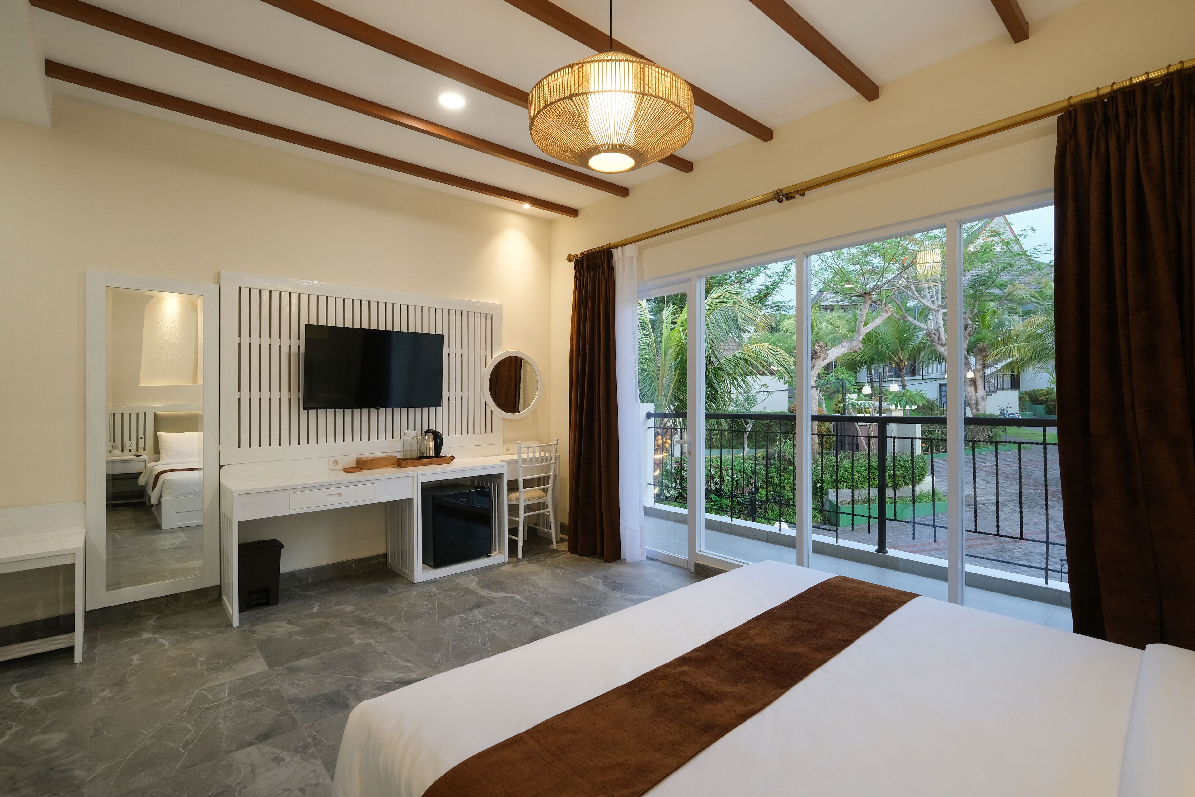 A hotel room with a bed, a desk, a TV, and a balcony with a view of palm trees.