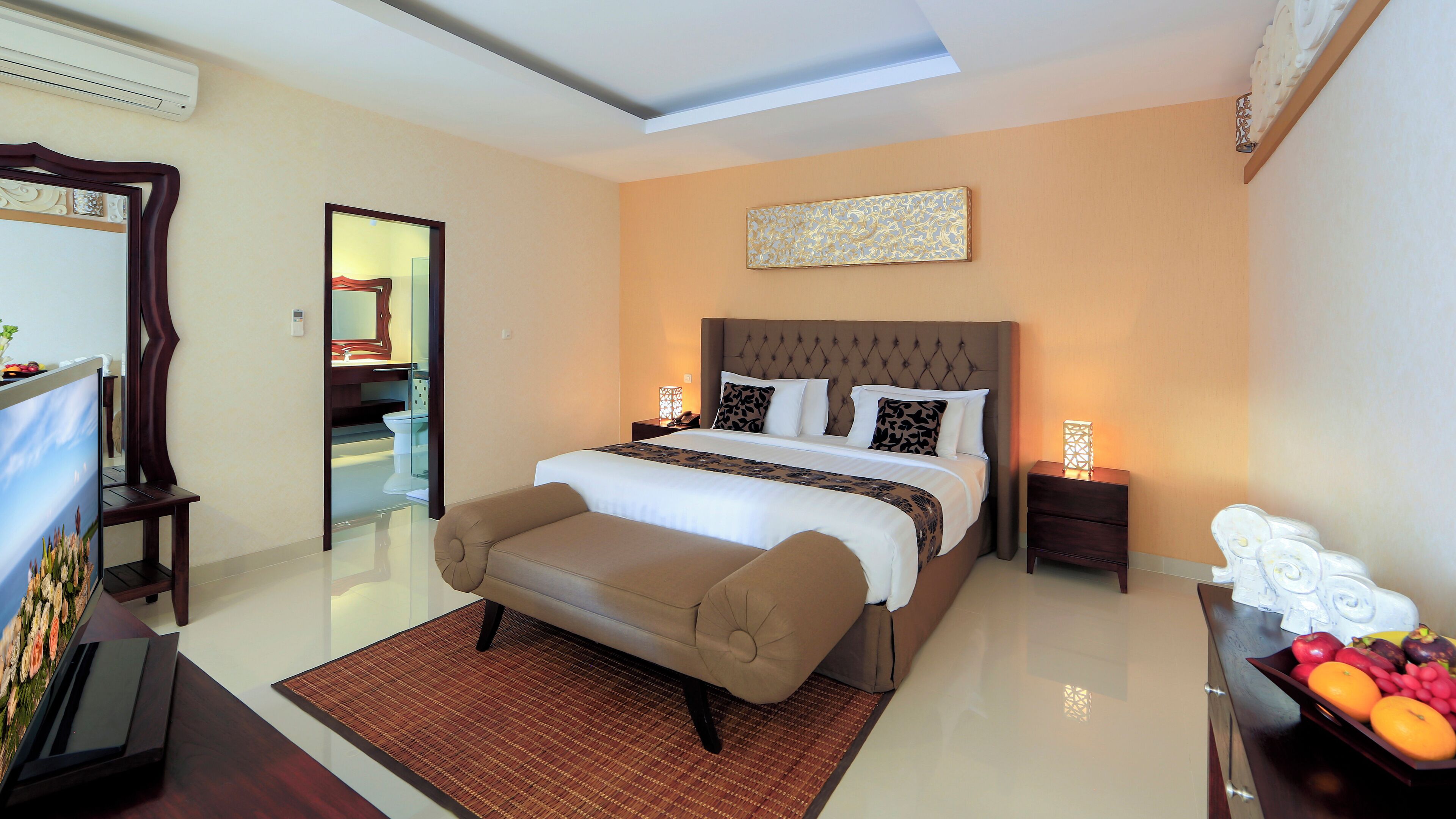 A modern hotel room with a large bed, a sitting area, a desk with a computer, and a small side table with a lamp.