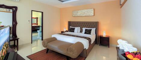 A modern hotel room with a large bed, a sitting area, a desk with a computer, and a small side table with a lamp.