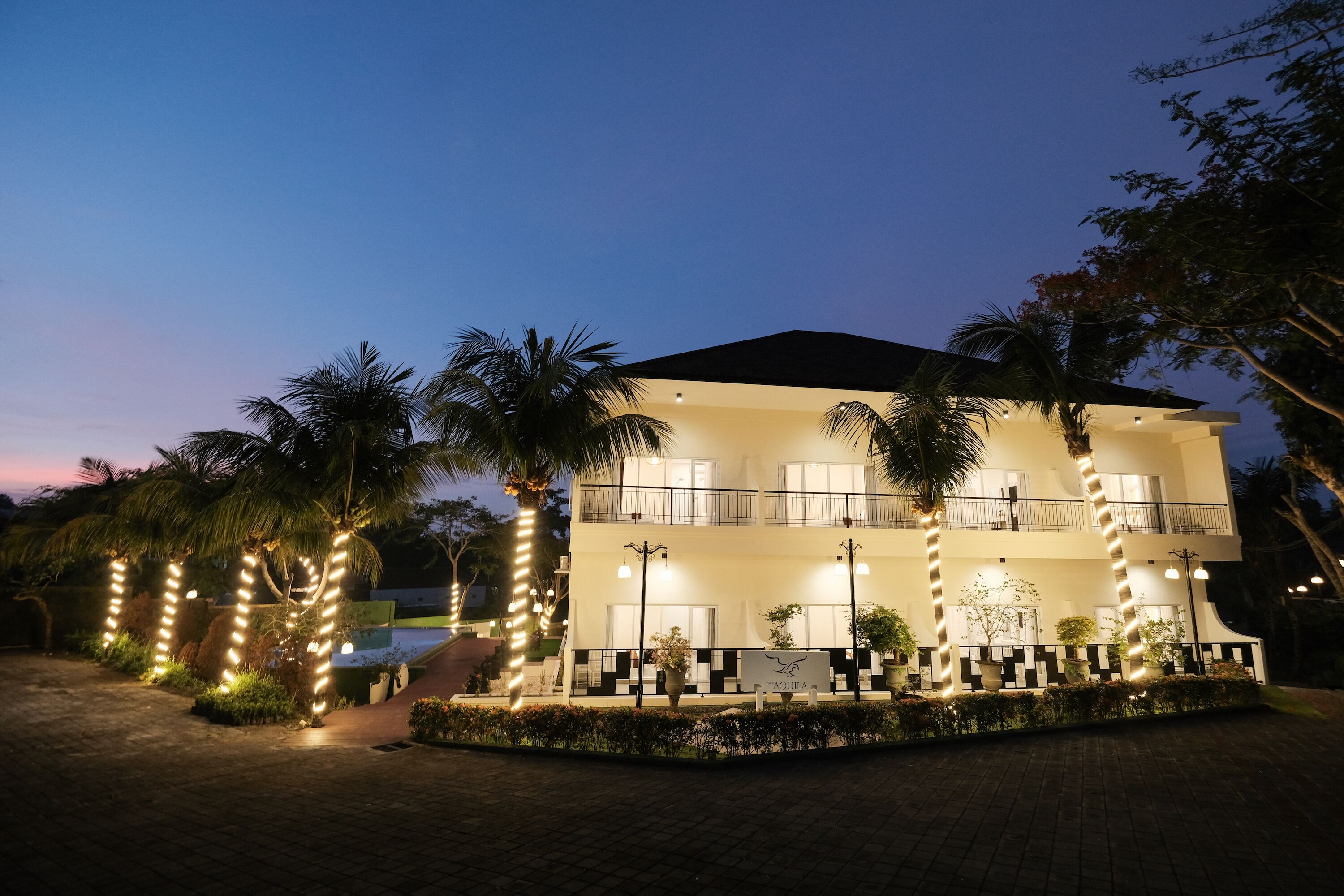 A two-story hotel with a balcony on the second floor, surrounded by palm trees and decorative lights.