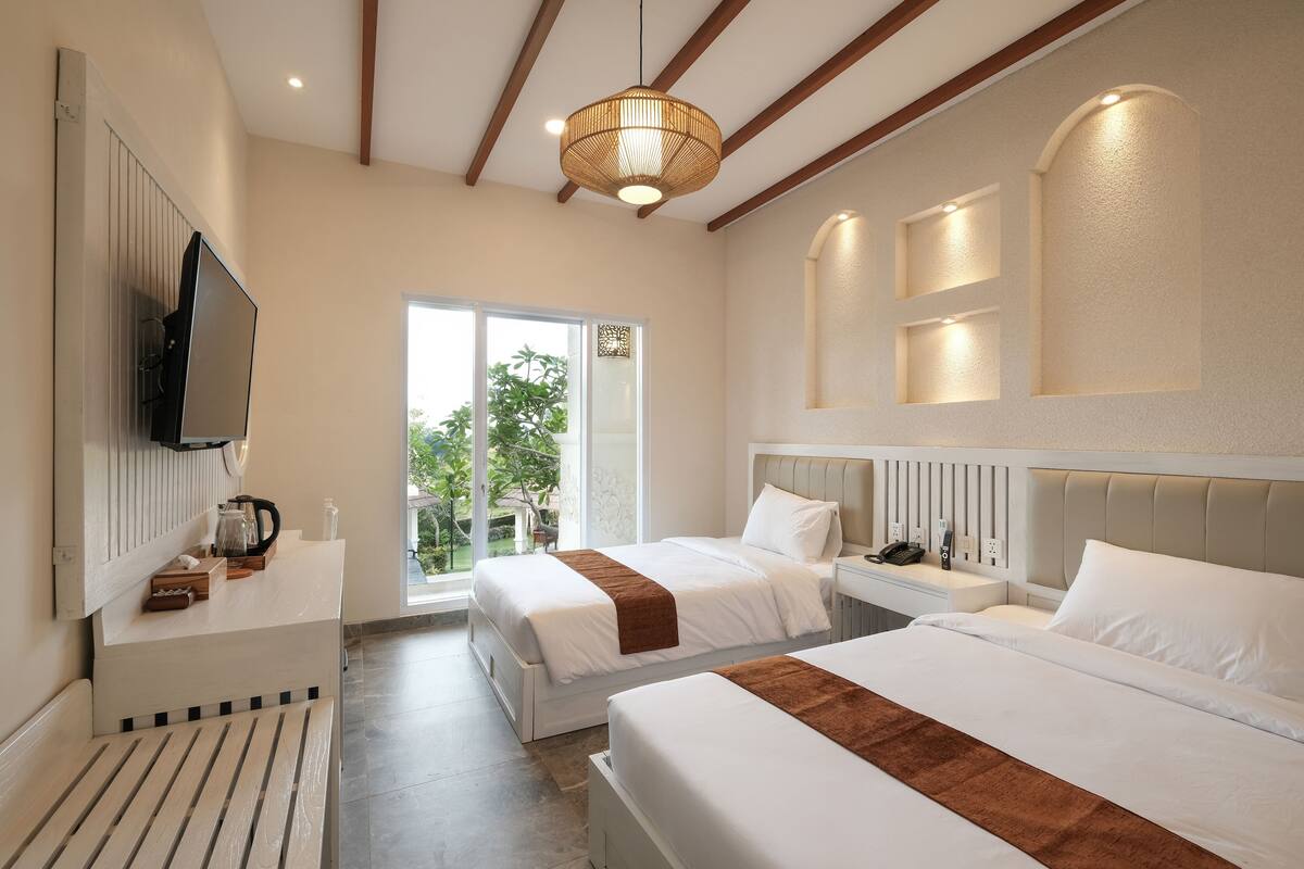 Habitación de hotel con dos camas, un televisor montado en la pared, una mesita con una tetera y vistas al exterior por una puerta corrediza.