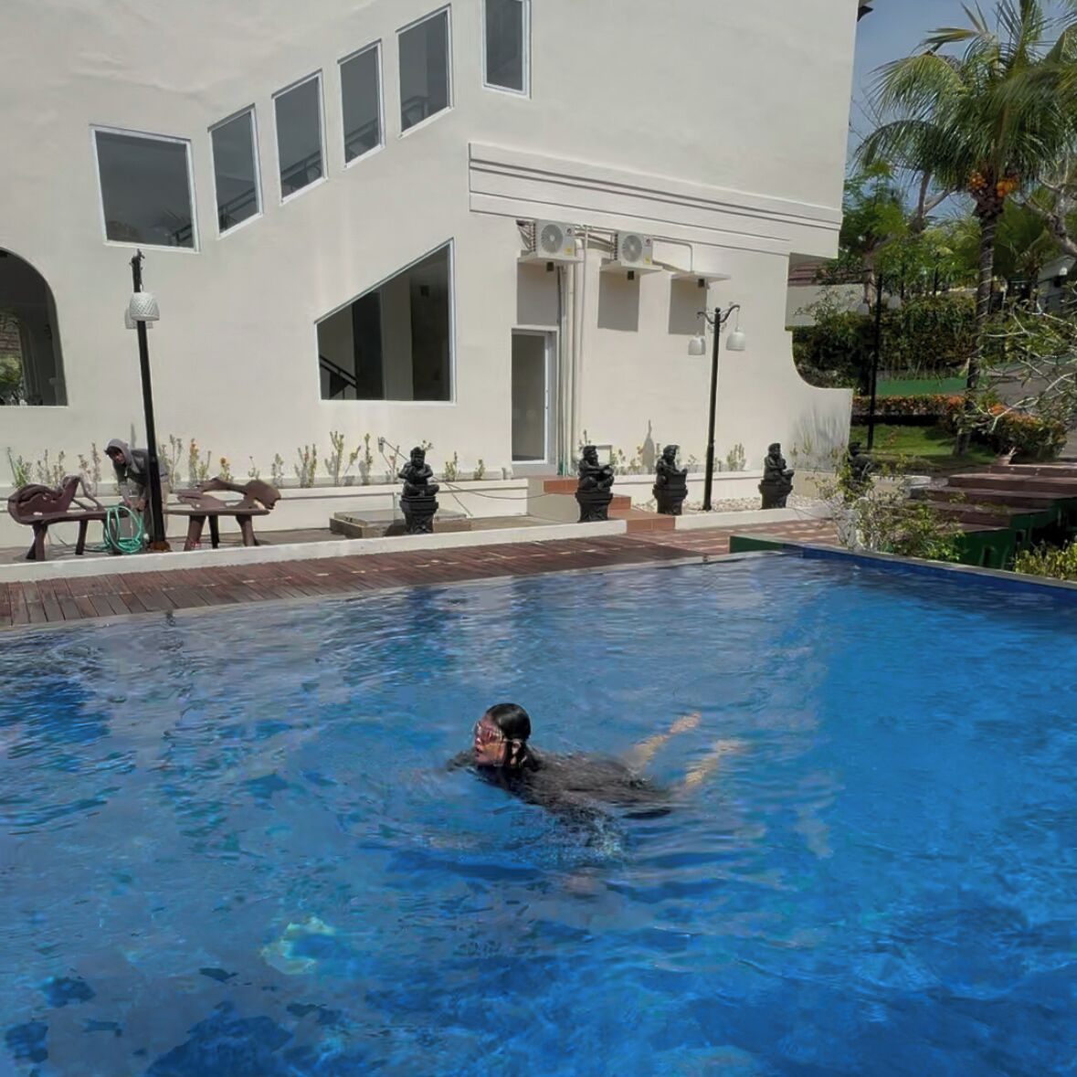 A swimming pool with a person swimming, a white building with multiple windows, and outdoor seating with statues.