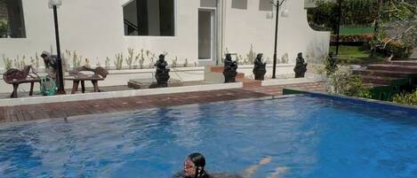 A swimming pool with a person swimming, a white building with multiple windows, and outdoor seating with statues.