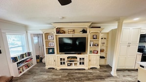 A living room with a large TV, wooden cabinets, and a bookshelf.
