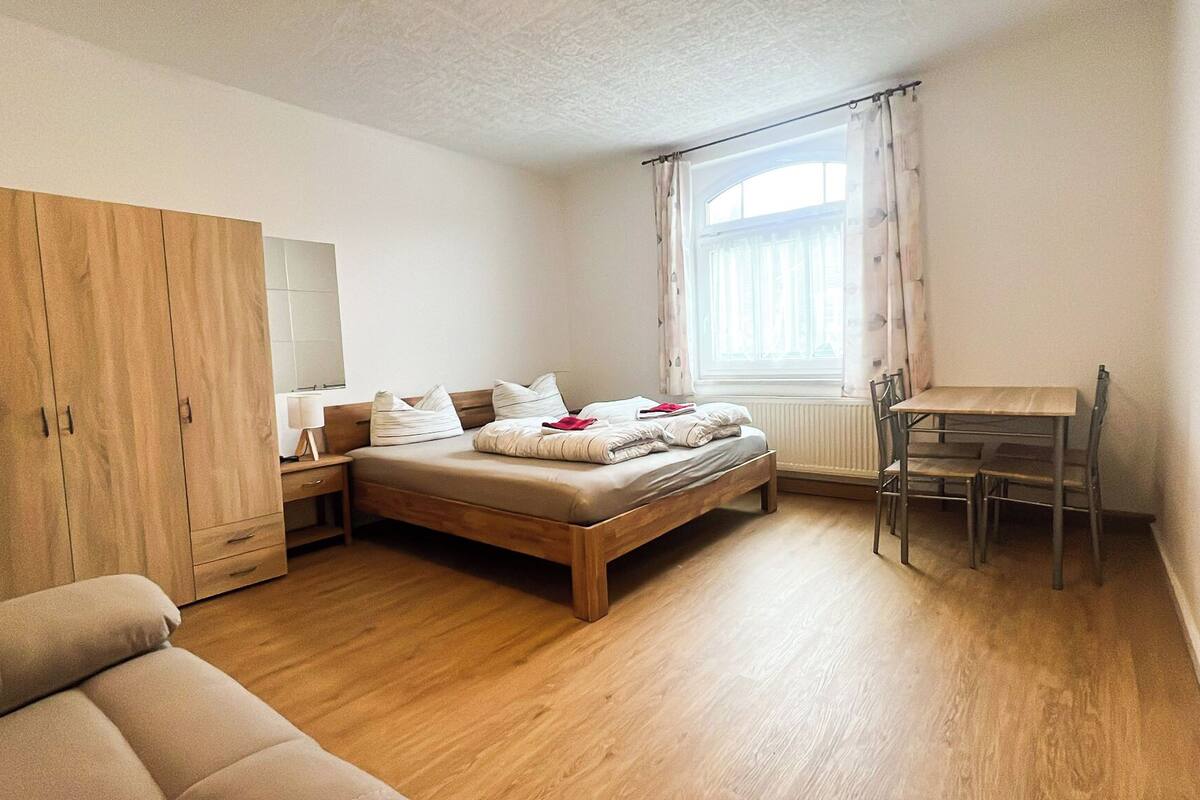 A bedroom with a wooden bed, a wooden wardrobe, a desk with a chair, and a window with curtains.