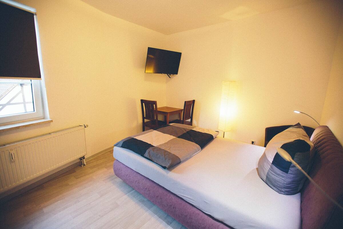 A bedroom with a bed, a TV mounted on the wall, a window, a chair, and a bedside table.