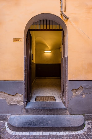 Un portail en arc donne accès à un espace intérieur faiblement éclairé, où l’on aperçoit un escalier et une enseigne indiquant « Scala G ».