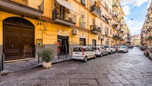 Eine enge Kopfsteinpflasterstraße, gesäumt von mehrstöckigen Gebäuden, parkenden Autos und einem Geschäft mit sichtbarer Schaufensterreklame.