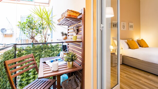 Ein Balkon mit einem Holztisch, Stühlen und Topfpflanzen. Durch eine offene Tür ist ein Bett mit weißer Bettwäsche und gelben Kissen zu sehen.