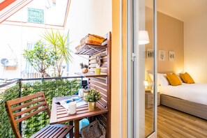Ein Balkon mit einem Holztisch, Stühlen und Topfpflanzen. Durch eine offene Tür ist ein Bett mit weißer Bettwäsche und gelben Kissen zu sehen.
