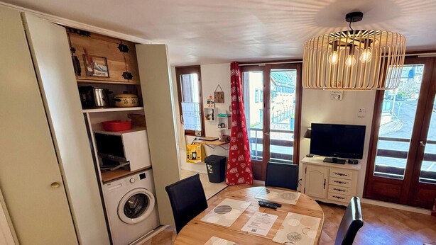 Un espacio habitable funcional con comedor, lavadora, televisor y vistas a través de un ventanal amplio.