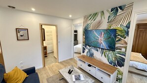 Una sala moderna con un televisor de pantalla plana montado en una pared decorada con un mural de hojas tropicales, un sofá, una mesa de centro con mandos a distancia y un pasillo que conduce a un dormitorio.