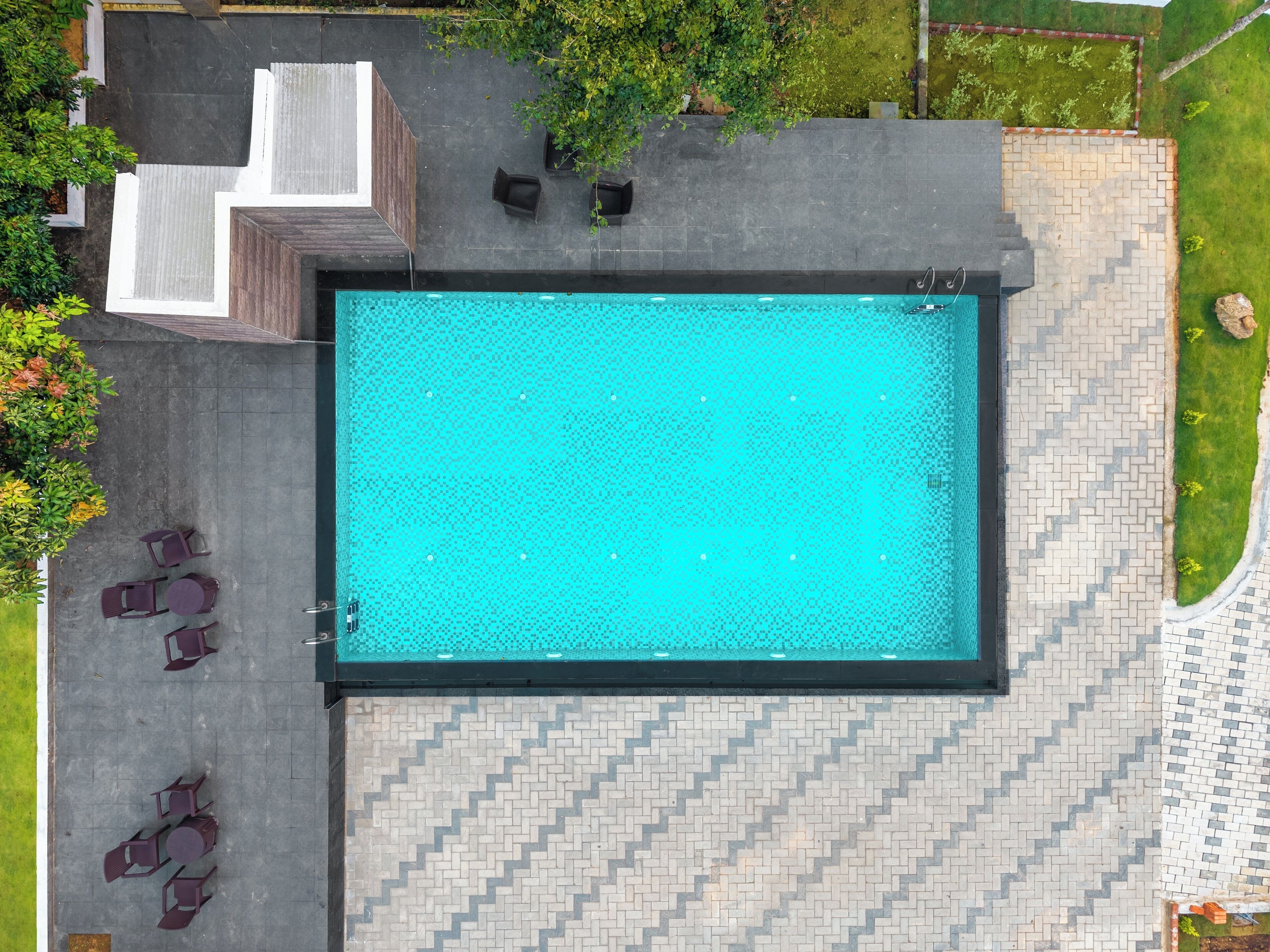 Vue aérienne d’une piscine dont le bord est revêtu de carreaux, entourée d’un espace de détente extérieur et de verdure.