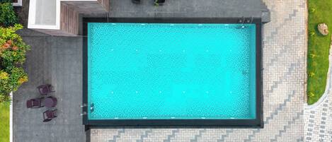 Vue aérienne d’une piscine dont le bord est revêtu de carreaux, entourée d’un espace de détente extérieur et de verdure.