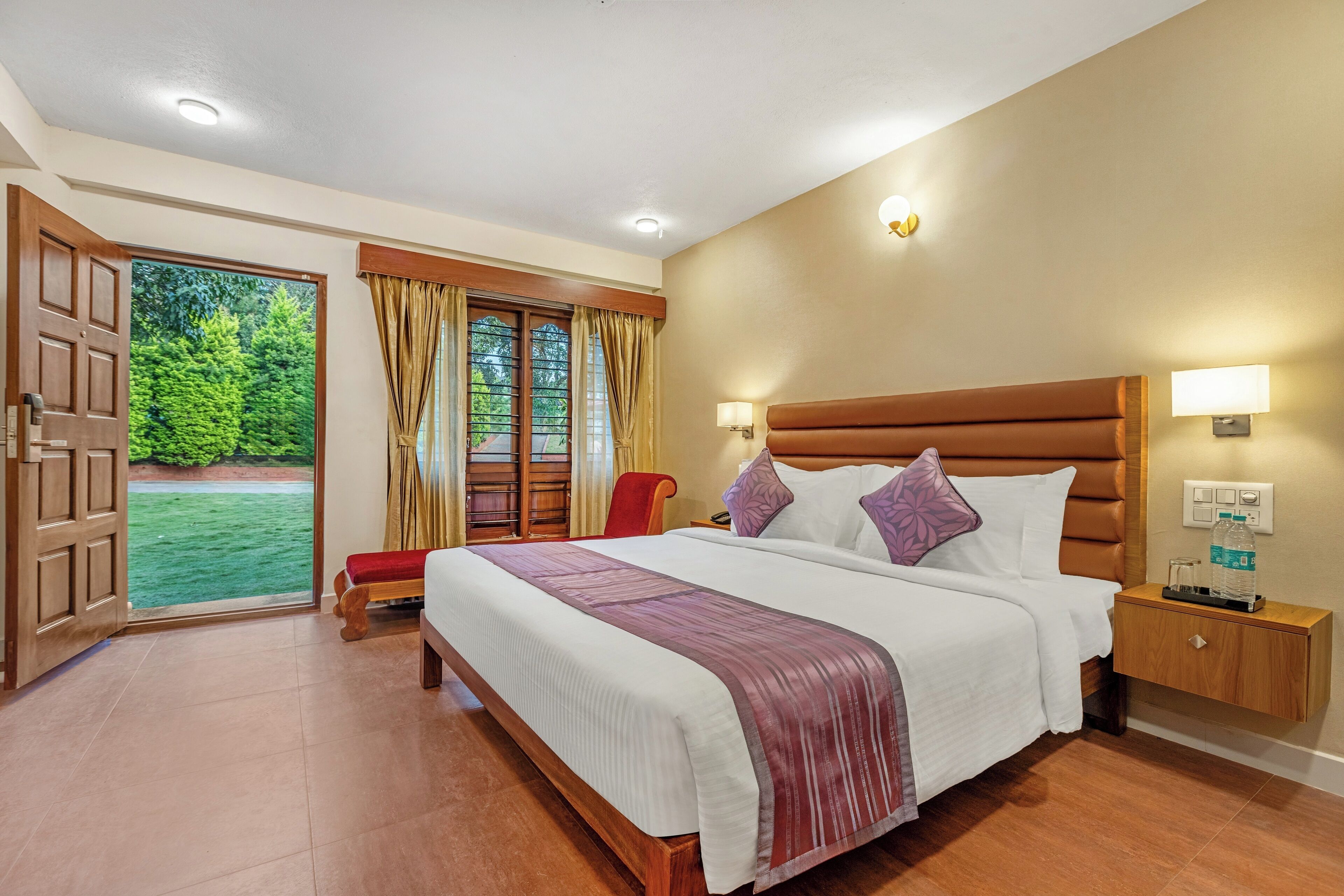 A spacious bedroom with a large bed, a wooden nightstand, a red chair, and a view of greenery through a sliding door.