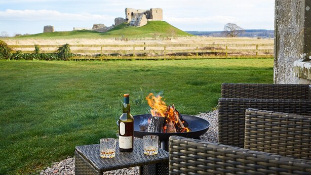 A patio with wicker furniture, a fire pit, and a bottle of wine. In the background, there is a grassy field and a castle ruin. - Old Duffus Farmhouse on Scottish Estate - stunning castle views (Elgin)