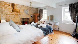 A bedroom with a stone wall, a bed with white linens, a wooden dresser, a vanity with a mirror, and a window with curtains. - Beautiful characterful cottage,family and dog friendly,quiet,close to amenities  (Harrogate)