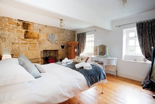 Een slaapkamer met een stenen muur, een bed met witte lakens, een houten dressoir, een kaptafel met spiegel en een raam met gordijnen.
