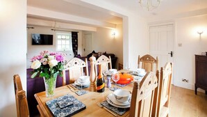A dining area with a wooden table set for four, surrounded by wooden chairs. A vase with flowers is on the table, and a television is mounted on the wall in the background. - Beautiful characterful cottage,family and dog friendly,quiet,close to amenities  (Harrogate)