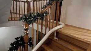 Eine hölzerne Treppe mit Handlauf, verziert mit Grünpflanzen und Lichtern, die zu einer oberen Etage führt.