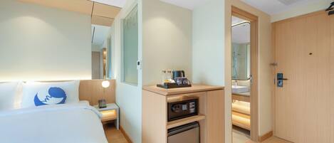 A hotel room with a bed, a desk, a mini-fridge, a mirror, and a door leading to a bathroom.