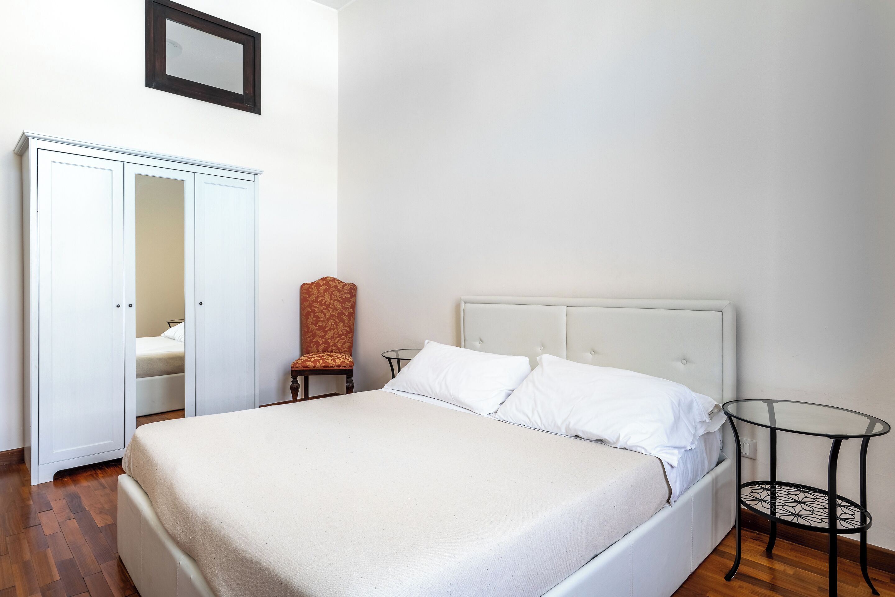 A neatly arranged bedroom with a bed, a wardrobe, a chair, and a small table.