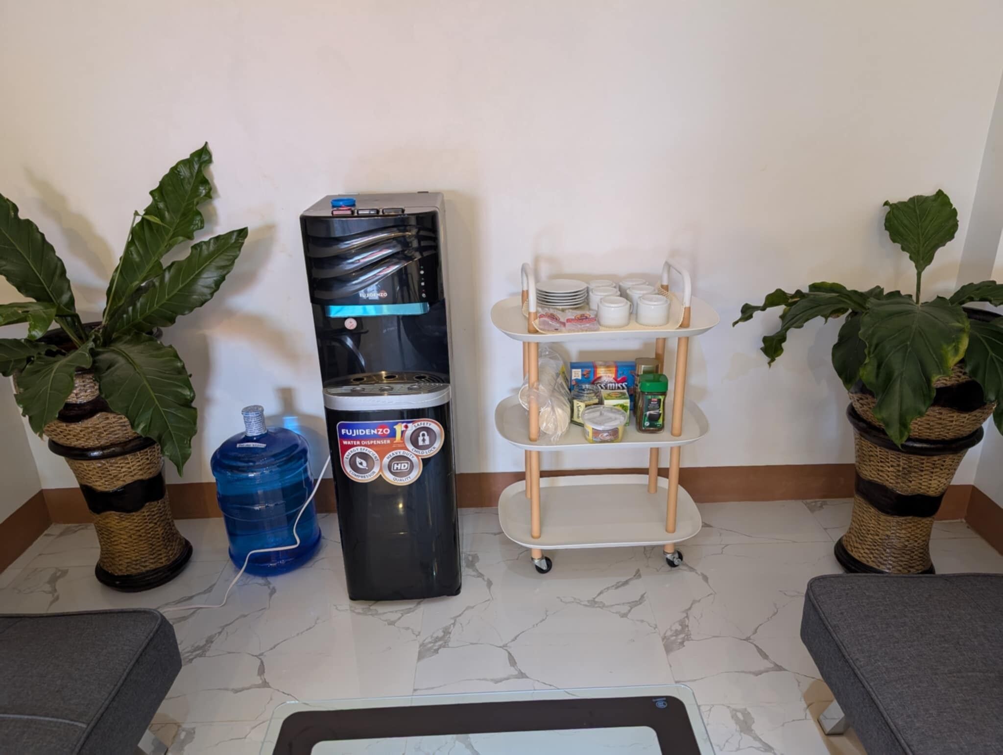A water dispenser, a small cart with dishes, and two potted plants.