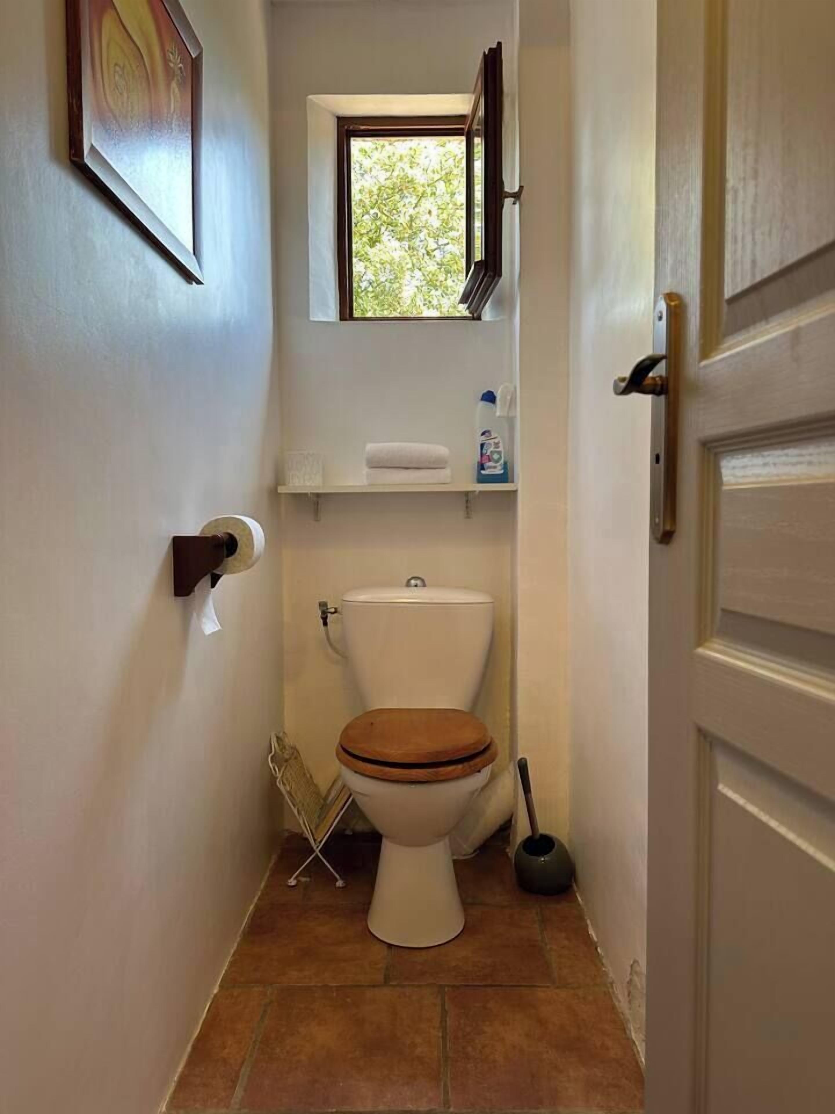 A small bathroom with a toilet, a window, a shelf with toiletries, and a door.
