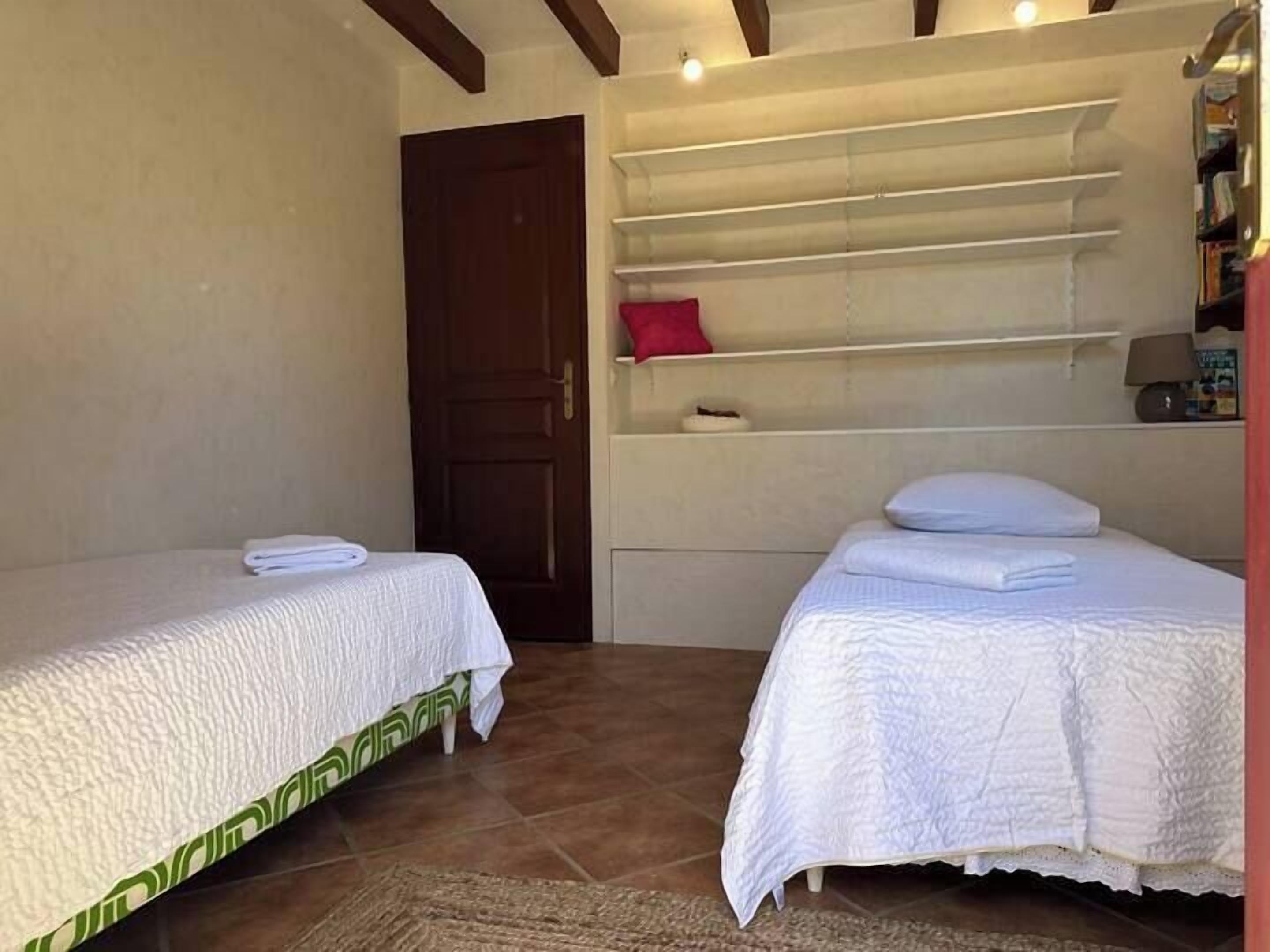 A room with two beds, a wooden door, and a shelf with a red pillow.