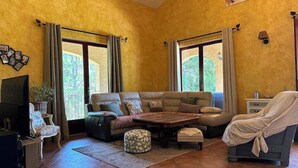 A living room with a sofa, ottoman, and a coffee table. There are two windows with curtains, a TV, and a lamp on the wall. - Beautiful Cevennes farmhouse with swimming pool (Corbès)