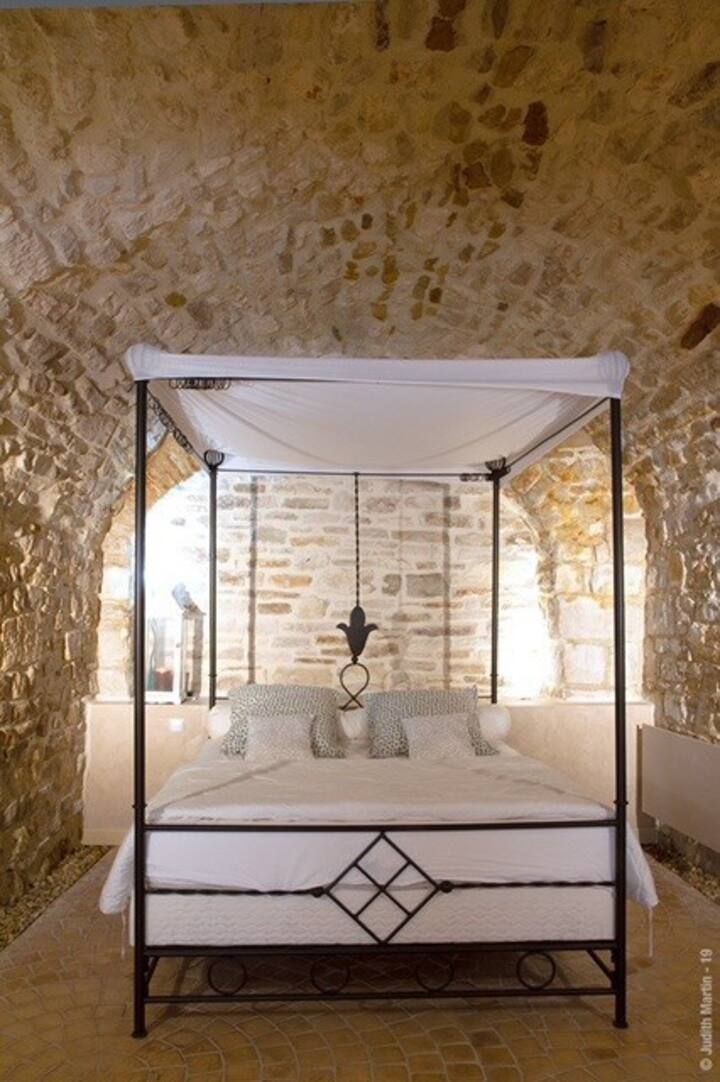 Een bed met hemelbed onder een hemelgordijn in een kamer met stenen muren en een rustieke sfeer.