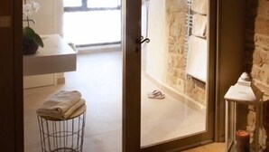 Puerta abierta que da a una habitación con pared de piedra, una ventana y una pequeña mesa con una toalla.