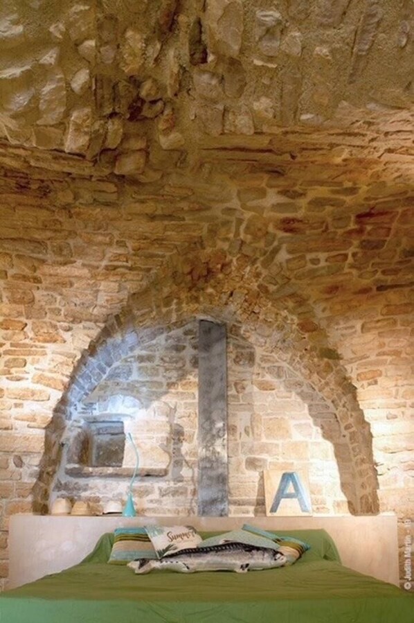 Habitación de paredes de piedra con una cama, un pequeño estante con una lámpara y una letra 'A' en la pared.