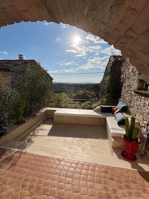 Une terrasse extérieure avec un banc en pierre, un cactus en pot rouge, et une vue sur un bâtiment en pierre et un paysage environnant.