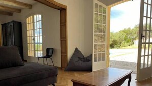 A spacious living room with a dark grey sofa, a wooden coffee table, and a black chair. A large window and a door with glass panels provide views of the outdoors. - L'oustal de cigales, panoramic view (Lédignan)