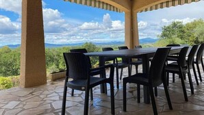A covered outdoor seating area with a stone floor and a table surrounded by chairs, overlooking a mountainous landscape. - L'oustal de cigales, panoramic view (Lédignan)