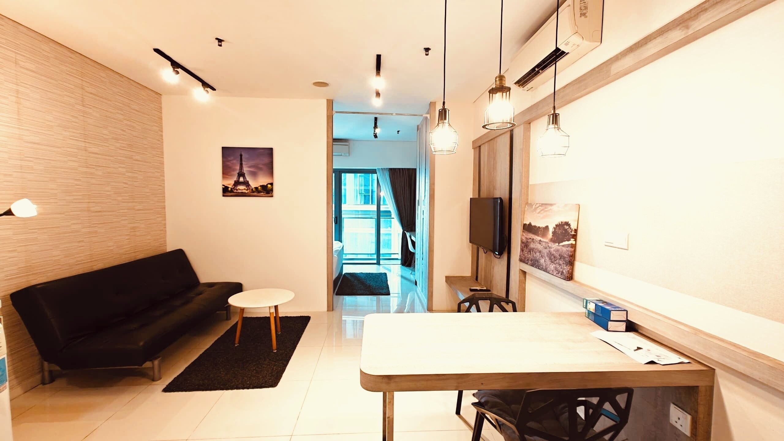 A modern living room with a black sofa, a small round table, and a wooden dining table. There are pendant lights above the dining table and a large mirror on the wall.