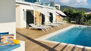 Un área de piscina moderna al aire libre con terraza de madera, sillas de descanso y vistas a las montañas.